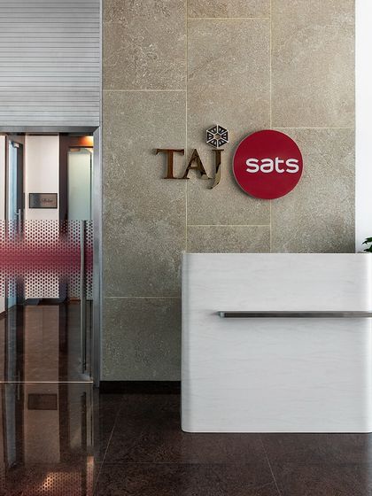 The reception desk at 'The High-Flying Office', set against a textured stone wall. The design is clean and minimalist, reflecting the professional and modern identity of the TajSATS brand.