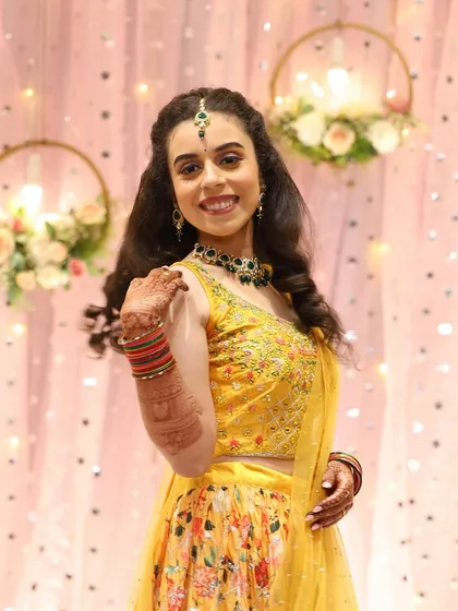 The bride looking radiant in her yellow floral lehenga for the sangeet. The makeup is fresh and bright, with a pop of color from the green jewelry.