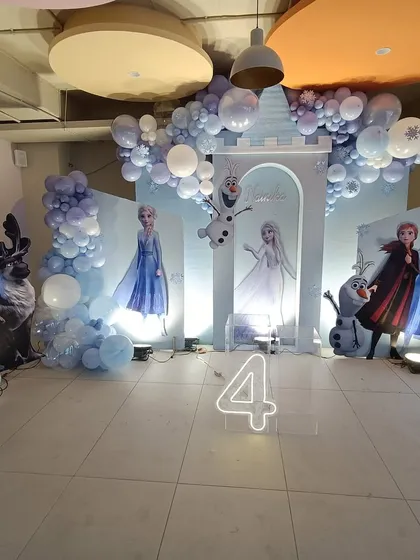 A full view of our enchanting Frozen-themed party for a fourth birthday. The scene is complete with character cutouts of Elsa, Anna, and Olaf, set against an ice castle backdrop and a cascade of blue, purple, and white balloons.