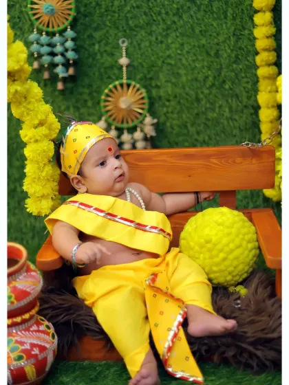 An alert baby dressed in a yellow Krishna costume sits on a miniature swing decorated with marigold garlands. This setup creates a playful and festive atmosphere for the photoshoot.
