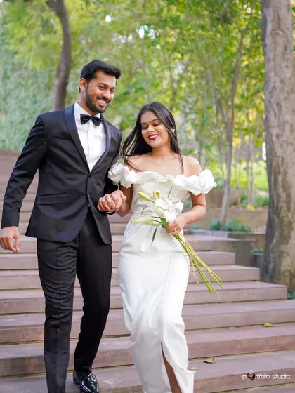 A happy couple, dressed in elegant formal wear, descends a staircase in a sunlit garden. This image showcases a classic and joyful pre-wedding style, capturing candid smiles and movement.