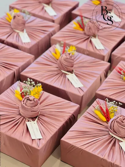 A batch of gifts elegantly wrapped in dusty rose fabric. Each gift is topped with a fabric rose knot and a small sprig of colorful dried flowers, creating a cohesive and beautiful presentation for a special event.