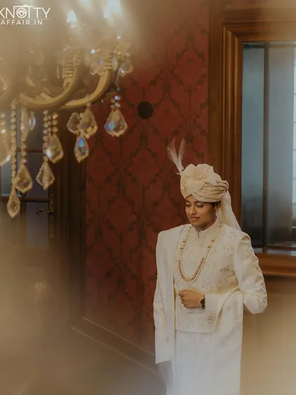 A portrait of the groom in his elegant white sherwani, captured through a soft focus foreground. The lighting and composition give this image an artistic, dreamlike quality.