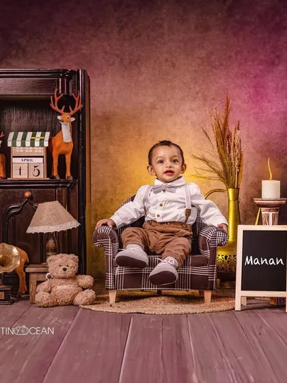 This rustic, woodland-themed studio setup is perfect for a classic toddler boy portrait. Dressed in a smart shirt and suspenders, he sits confidently in a scene filled with warm, earthy tones, deer figures, and soft candlelight.