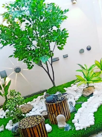A small, custom garden landscape created for an indoor space. It features an artificial tree, various small plants, and decorative elements like white pebbles and wood stumps.