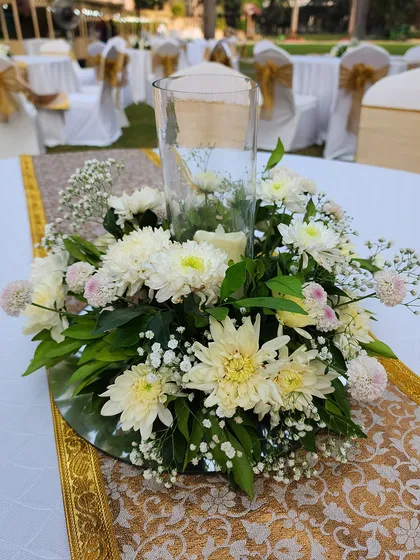 A lovely low centerpiece for a daytime event, featuring white chrysanthemums and baby's breath around a glass candle holder. The arrangement sits on a mirrored base to reflect the light.