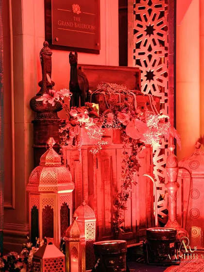 A corner vignette styled to look like a treasure trove, with antique-style lanterns, wooden crates, and floral arrangements, all bathed in a warm red light.