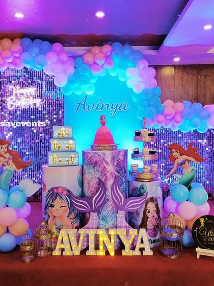 A wider view of the Mermaid-themed birthday decor, showcasing the full stage with its shimmering backdrop, themed plinths, and colorful balloon arch.