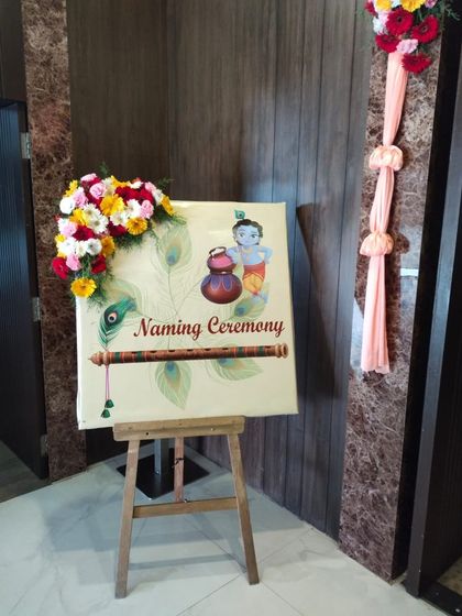A custom welcome sign for a naming ceremony featuring a cute baby Krishna illustration. The sign is placed on a wooden easel and decorated with fresh flowers.