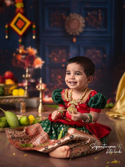 A beautiful, smiling toddler girl in a traditional South Indian dress, surrounded by festive decorations.