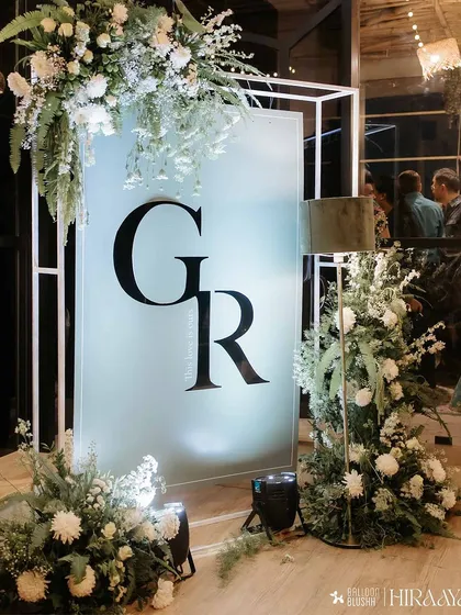 A detailed shot of the custom monogrammed light box. Framed by asymmetrical arrangements of white chrysanthemums, ferns, and other greenery, it serves as a modern and personalized welcome statement for the couple's celebration.