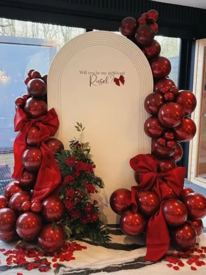 A grand romantic gesture. This "Will you be my girlfriend?" proposal setup features a stunning arrangement of red chrome balloons, large bows, and scattered rose petals.