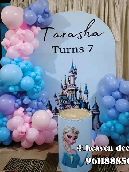 A simple yet charming Frozen-themed setup for a 7th birthday. The decor features an arched backdrop with a castle graphic, a character-printed plinth, and a balloon arrangement in pink, blue, and purple.