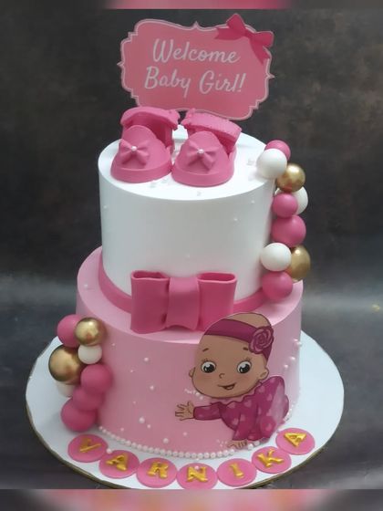 A beautiful two-tier welcome cake for a baby girl. This fresh pineapple cake is decorated in pink and white with fondant baby shoes and a big bow.