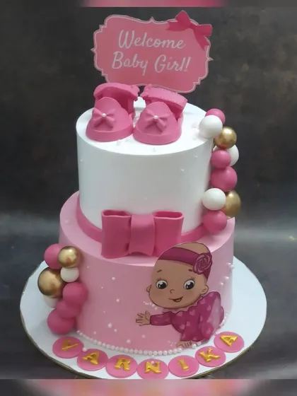 A beautiful two-tier welcome cake for a baby girl. This fresh pineapple cake is decorated in pink and white with fondant baby shoes and a big bow.
