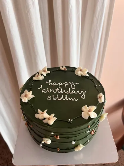 An elegant olive green cake with white piped flowers and gold sprinkle details. The horizontal lines on the side add a modern texture.