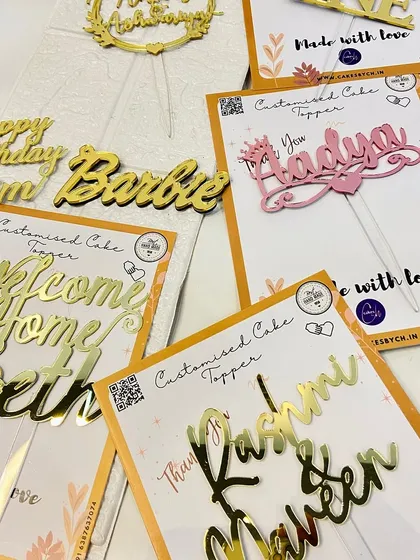 A collection of various toppers, including a pink "Aadya" and a gold "Barbie" name topper, showing the range of styles available.