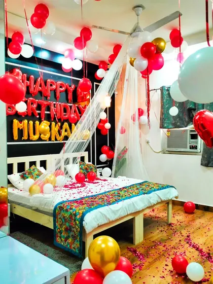 This birthday room decoration for 'Muskan' features a beautiful canopy with fairy lights, lots of red, white, and gold balloons, and a floor scattered with petals. It’s a very popular combination.
