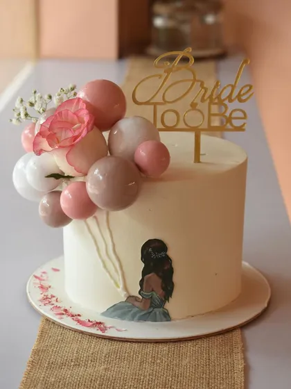Peach and prettiness for this bride-to-be. This cake features a hand-painted illustration and a whimsical balloon-like arrangement of spheres in shades of pink, complemented by a fresh flower and a gold topper.