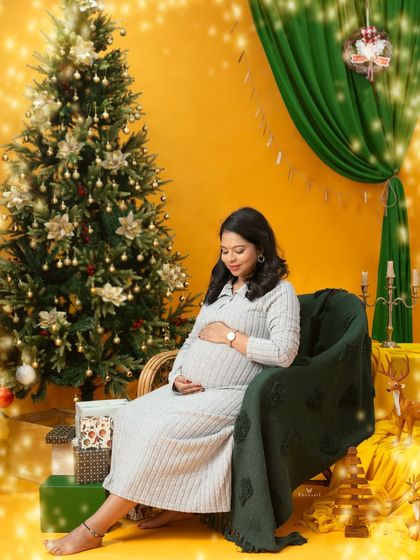 A cozy Christmas-themed maternity shoot. The mom-to-be is seated by a decorated tree, wearing a comfortable sweater dress for a warm and casual holiday portrait.