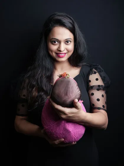 A beautiful portrait of a mother smiling as she holds her sleeping baby. The focus is entirely on the mother and her newborn against a classic dark background.