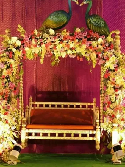 A beautiful jhula for a Janmashtami or Teej celebration. The swing is covered in a dense arrangement of colorful flowers, with two peacock statues sitting on top, creating a festive and traditional look.