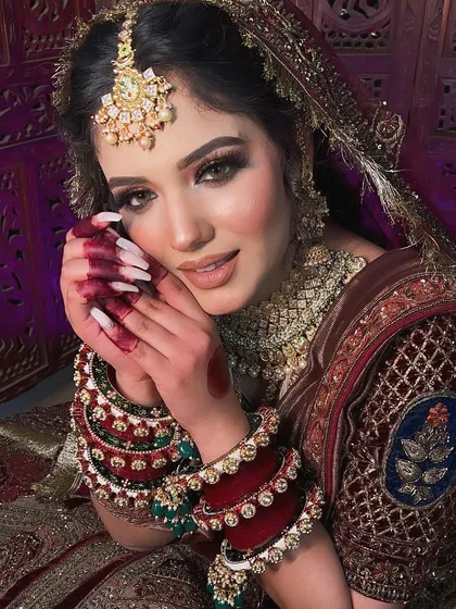 A soft, romantic bridal portrait. The focus is on the glowing skin and the beautiful expression of the bride, with her henna-adorned hands framing her face.