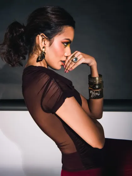 A profile shot that showcases the sheer fabric of the top and the elegant line of the model's back. The lighting is carefully placed to highlight the silhouette and the intricate details of the accessories, creating a sophisticated and moody fashion portrait.