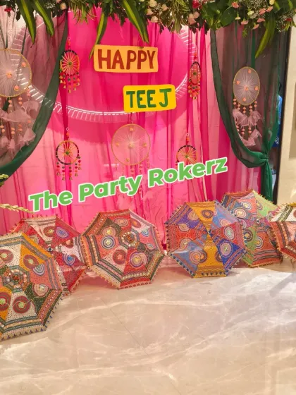 A festive Teej photo corner with a pink and green drape backdrop, decorated with dreamcatchers and colorful embroidered umbrellas.