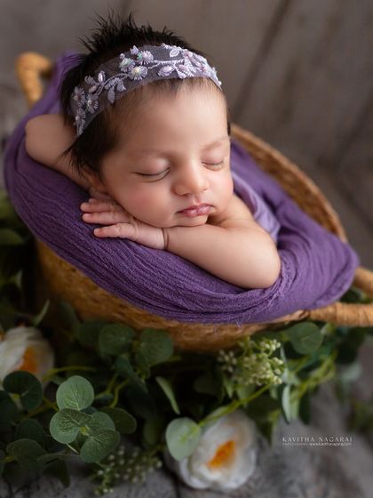 Three-week-old Samaya in the chin-on-hands pose, surrounded by greenery. The purple wrap and delicate headband add a touch of regal elegance.