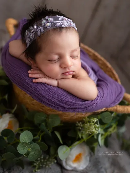 Three-week-old Samaya in the chin-on-hands pose, surrounded by greenery. The purple wrap and delicate headband add a touch of regal elegance.
