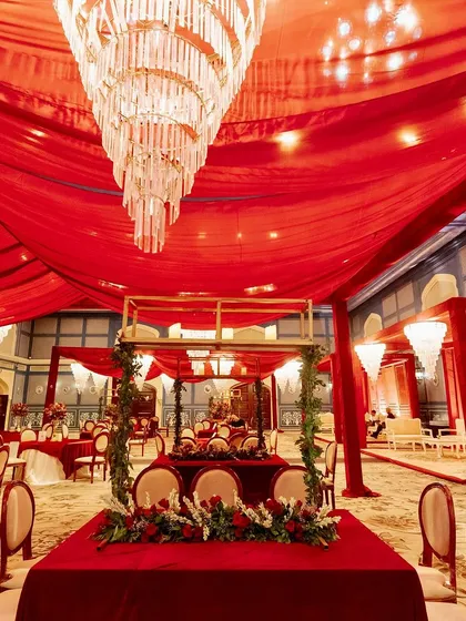 An indoor wedding setup where the entire ceiling is draped in rich red fabric, with a massive crystal chandelier as the centerpiece, creating a regal and opulent atmosphere.