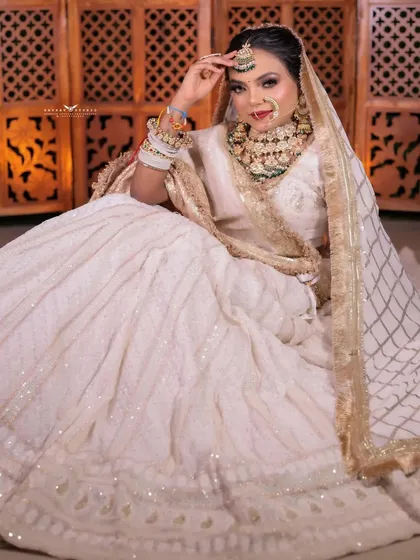 A full seated portrait, the white lehenga spread out beautifully against a traditional carved screen.