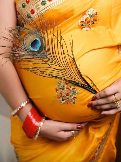 A close-up detail shot focusing on the baby bump, adorned with a peacock feather. This artistic photo highlights the texture of the yellow saree and the traditional red bangles, symbolizing beauty and new life.