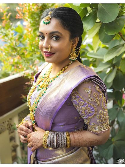 A stunning portrait of the bride, showcasing her beautiful lavender silk saree, intricate gold jewelry, and happy glow.