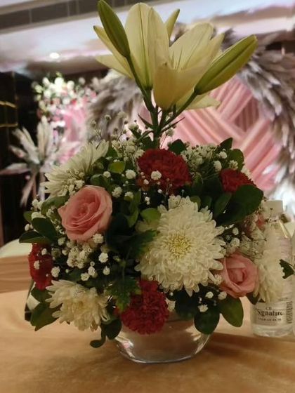 This is another example of a floral centerpiece for a guest table. The arrangement features a mix of lilies, roses, and chrysanthemums in a simple glass bowl, adding a fresh touch to the table.
