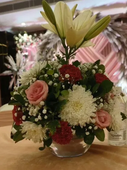 This is another example of a floral centerpiece for a guest table. The arrangement features a mix of lilies, roses, and chrysanthemums in a simple glass bowl, adding a fresh touch to the table.