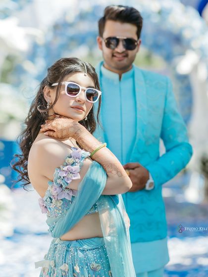 The bride strikes a cool pose in her sunglasses and beautiful blue lehenga, with her groom standing beside her, capturing their modern style at their Pushkar wedding.
