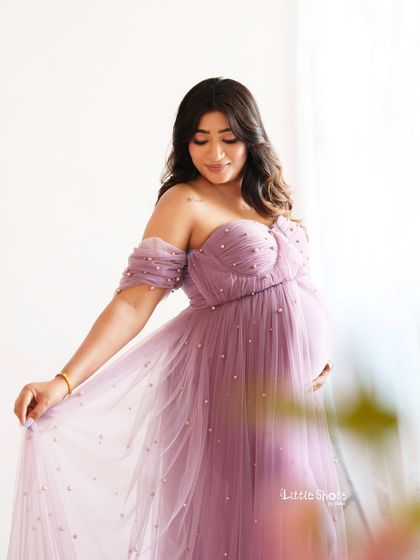 A graceful portrait of an expecting mother in a pearl-adorned lavender gown. She gently holds the flowing fabric of her dress, looking down with a peaceful expression.