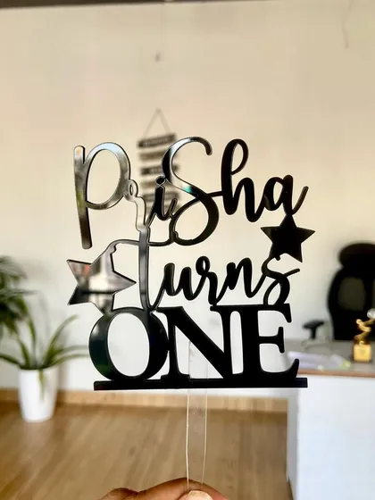 The "Prisha turns ONE" topper in black acrylic, held up to show its design clearly.