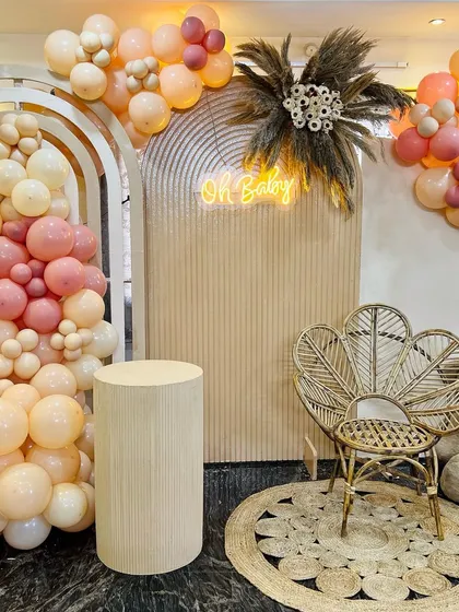 A warm and inviting boho baby shower setup. The decor includes neutral-toned balloons, a textured backdrop with a neon 'Oh Baby' sign, a peacock chair, and a cuddly teddy bear.