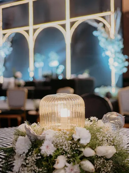 A centerpiece from the 'Love & Illusion' Sangeet. A modern ribbed glass lamp surrounded by baby's breath and silver-sprayed anthuriums, perfectly matching the chic, monochrome theme.