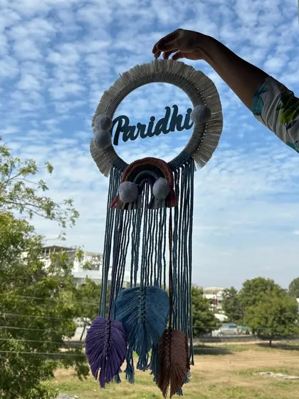 A beautiful shot of our custom macrame dreamcatcher for 'Paridhi' against the sky. The combination of the name, rainbow, and feathers makes it a whimsical and personal piece of art.