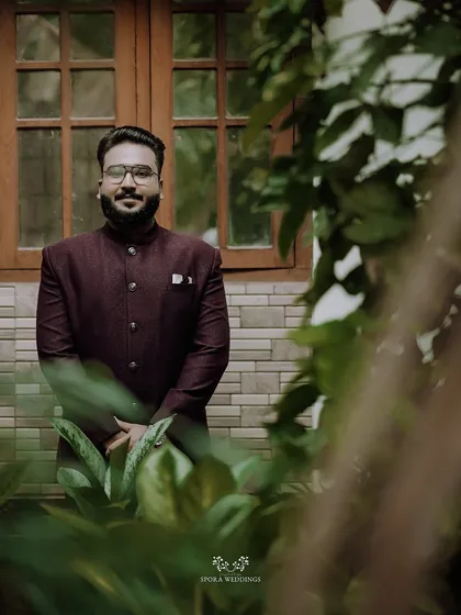 A handsome portrait of the groom-to-be in his stylish maroon bandhgala jacket, framed by lush greenery.