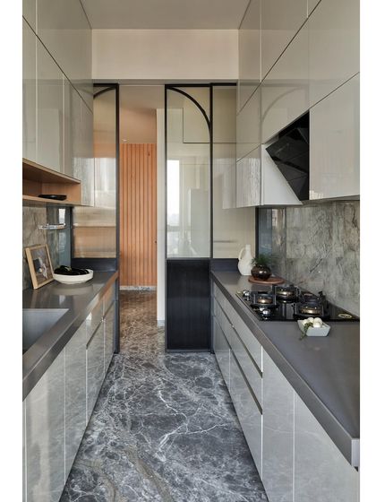 This sleek, modern galley kitchen features high-gloss grey cabinets and a dramatic marble floor. The design uses reflective surfaces to make the narrow space feel larger, while the glass partition maintains a connection to the rest of the home.