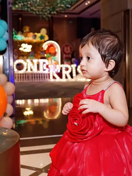A candid shot of a baby girl in a red dress looking at the party decorations, with the venue's ambiance reflected in the background.