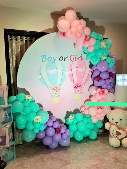 A "Boy or Girl" gender reveal party with a hot air balloon theme. The round backdrop is surrounded by teal, purple, and pink balloons, with "BABY" blocks and a teddy bear adding to the charm.