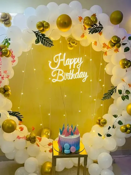 A simple and elegant birthday photo corner with a circular balloon arch in white and gold, a neon sign, and a plinth for the cake.