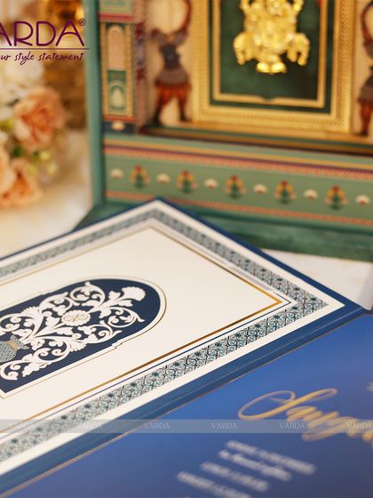 A close-up view of a bespoke wedding invitation suite. The design features intricate laser-cut patterns on a deep blue folder, which opens to reveal a Ganesha idol set within a miniature golden temple arch.
