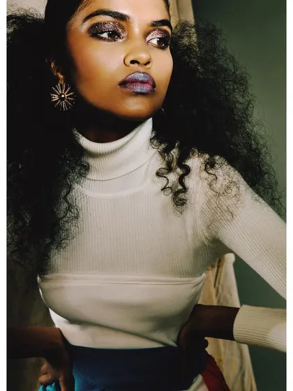 A close-up beauty shot of 'ANAINA,' showcasing glittery eye makeup and a metallic lip. The image highlights the collaboration between art direction and makeup artistry.